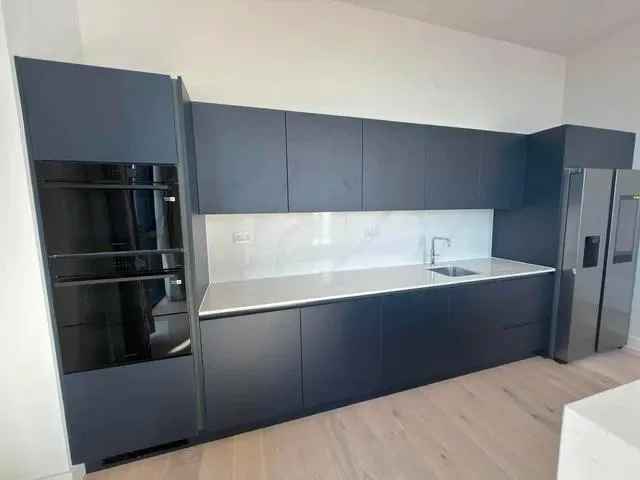 Dark wall and base cabinets with stacked ovens, a sink and a tall fridge freezer.